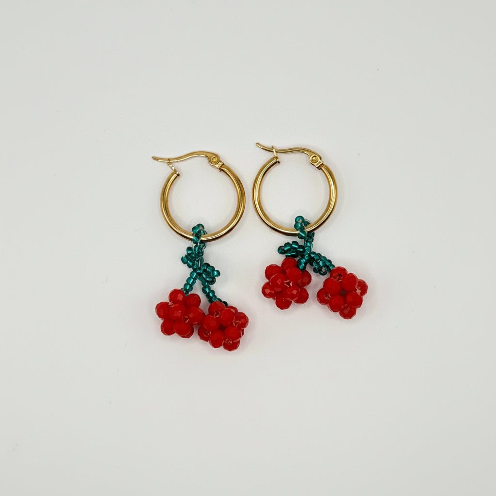 A pair of gold hoop earrings with dangling red cherry clusters and green stems, photographed on a plain white background.