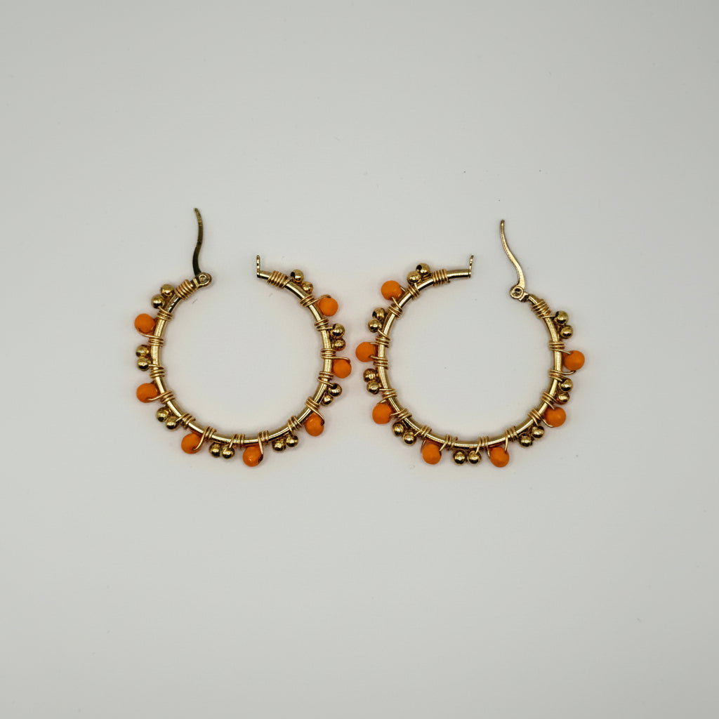 A pair of gold-tone hoop earrings decorated with small orange beads and metallic gold spacer details, displayed on a plain light gray background.