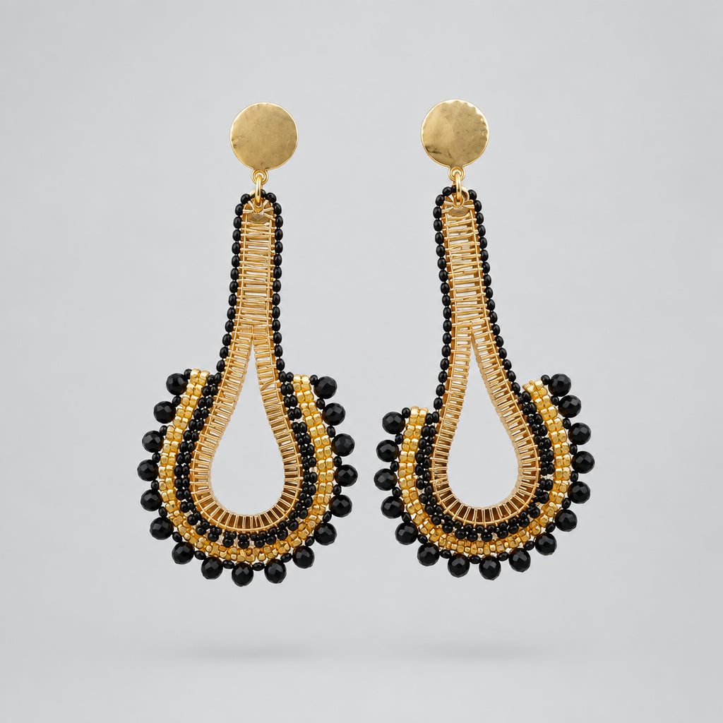 A pair of gold and black beaded teardrop dangle earrings on a white display card, set against a white textured background.