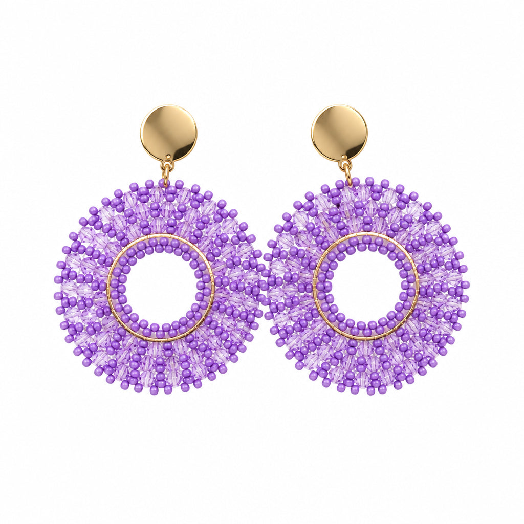 A pair of handmade beaded dangle earrings with purple seed beads in a circular sunburst pattern, featuring gold-toned metal studs and posts, displayed on a white jewelry stand.