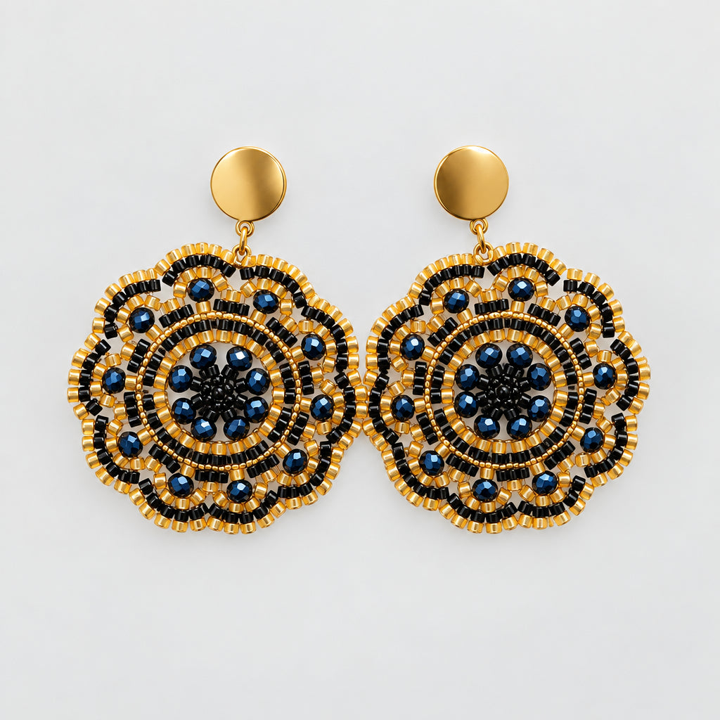 A pair of handmade circular drop earrings with intricate gold and black beaded patterns and dark blue center details, hanging from gold-toned posts on a clear display card against a white background.