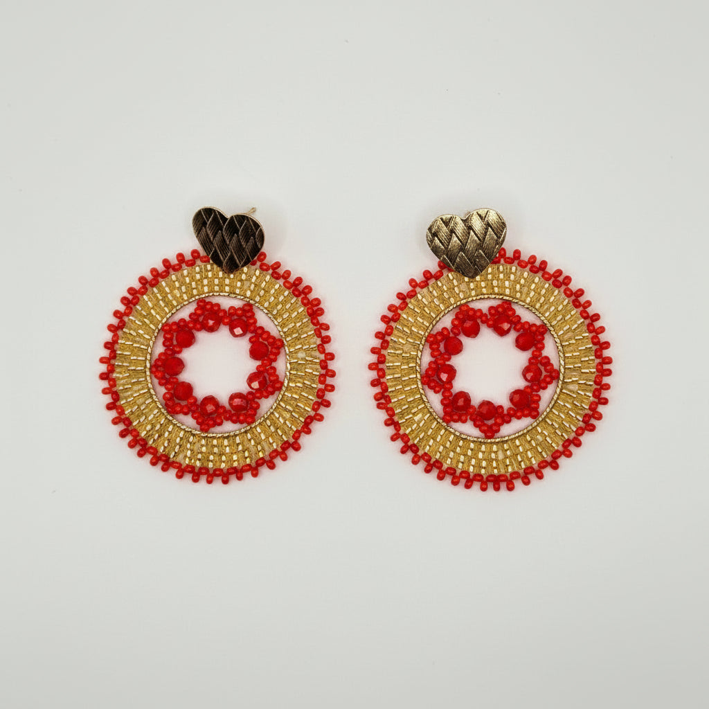 A pair of handmade circular beaded earrings with red and gold beadwork, a central red floral motif, and a small dark brown wooden heart stud at the top, displayed on a clean white background.