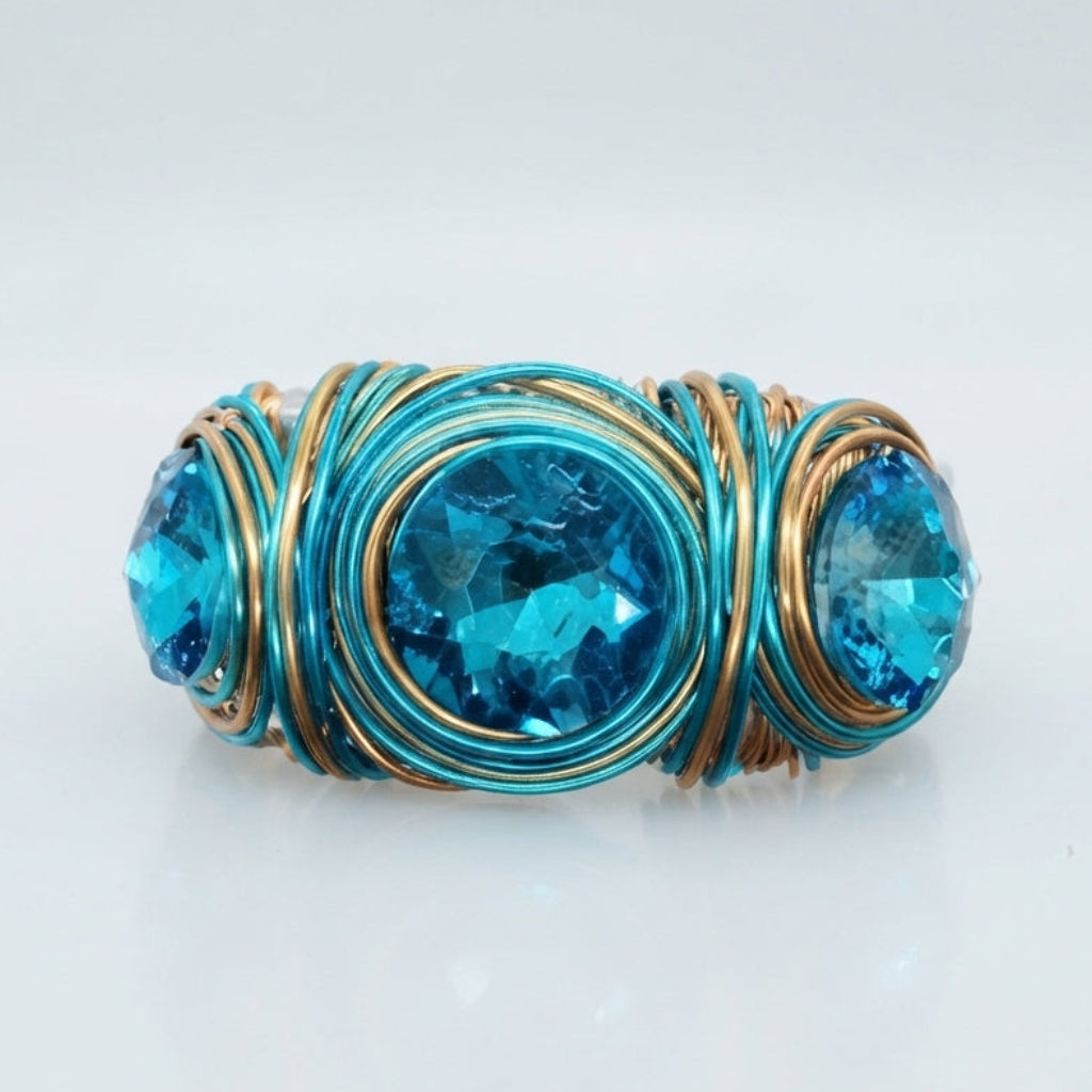 A handmade wire-wrapped ring featuring three oval-cut bright blue gemstones, set in a textured wrap of turquoise and gold-toned metal, shown on a clean white background.