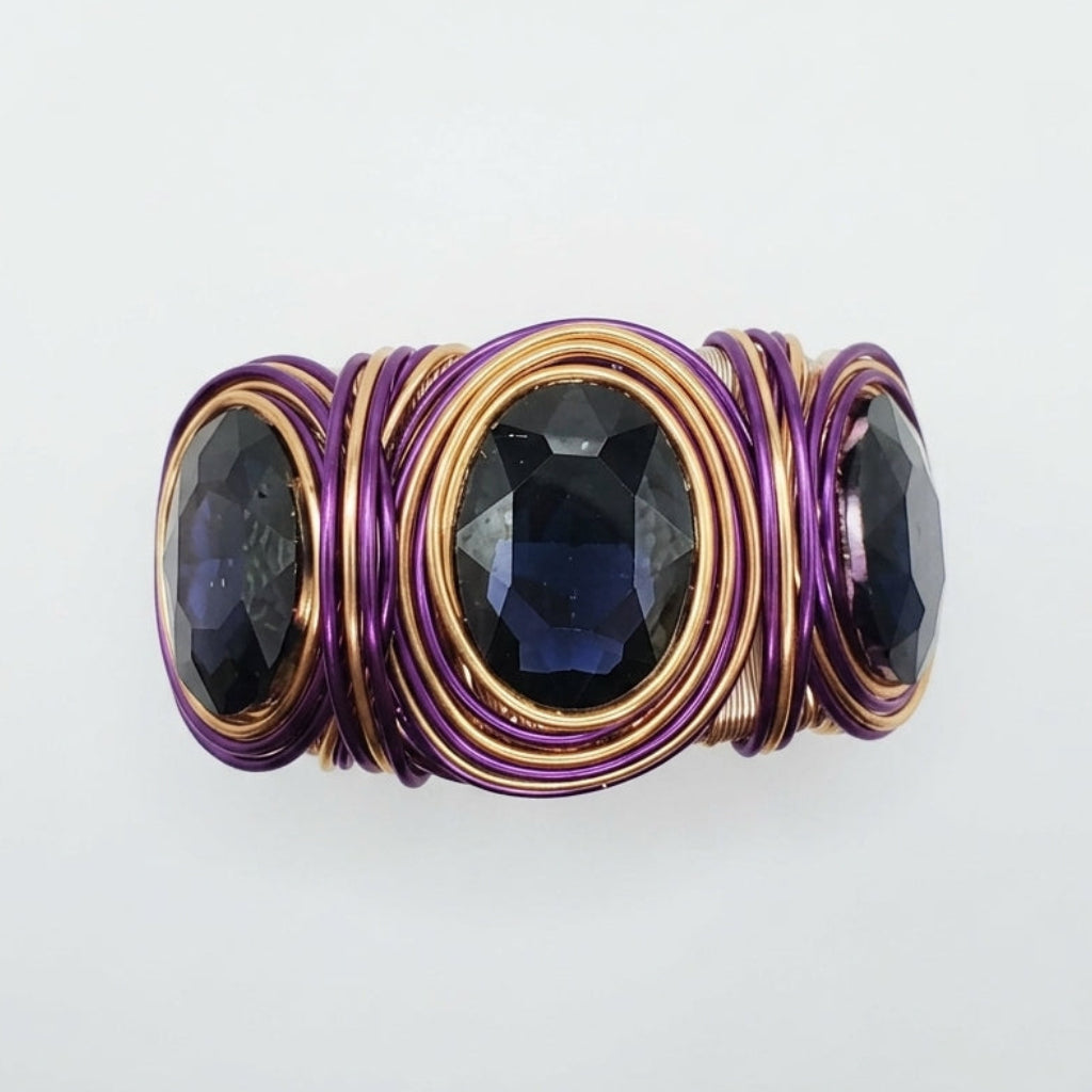 A handmade wire-wrapped ring featuring three oval faceted dark blue gemstones, wrapped in alternating layers of purple and gold-toned copper wire, set against a plain white background.