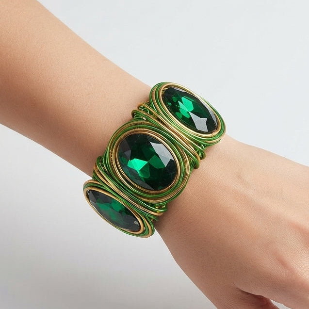 A close-up of a person's hand and wrist wearing a wide green and gold wrap bracelet with three large oval green gemstones, set against a clean white background.