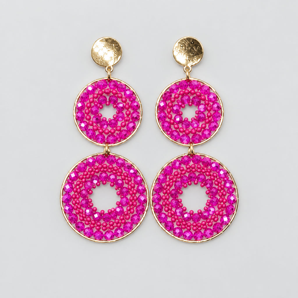 A pair of dangling earrings displayed on a white jewelry stand. Each earring has a small round gold stud at the top and a larger circular drop below covered in bright pink rhinestones with a small white center cutout.