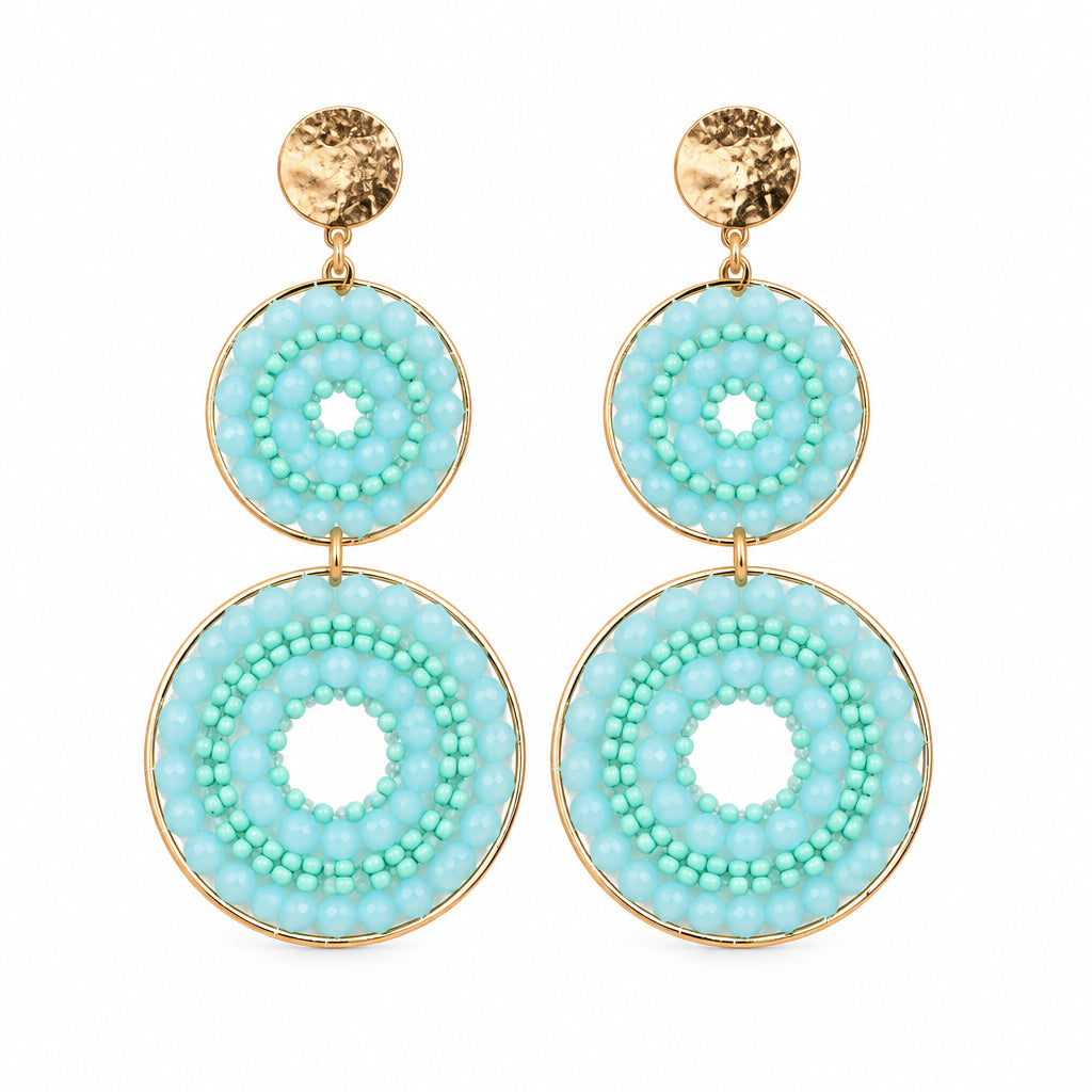 A pair of drop earrings with circular turquoise and white patterned discs on a white display card labeled STAINLESS STEEL, set against a white textured background.