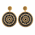 A pair of gold-tone dangle earrings with circular black and gold mandala-style patterns, displayed on a white jewelry stand.