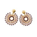 A pair of gold-toned dangle earrings with intricate circular beaded patterns in gold, white, and dark purple, displayed on a small white card against a white background.
