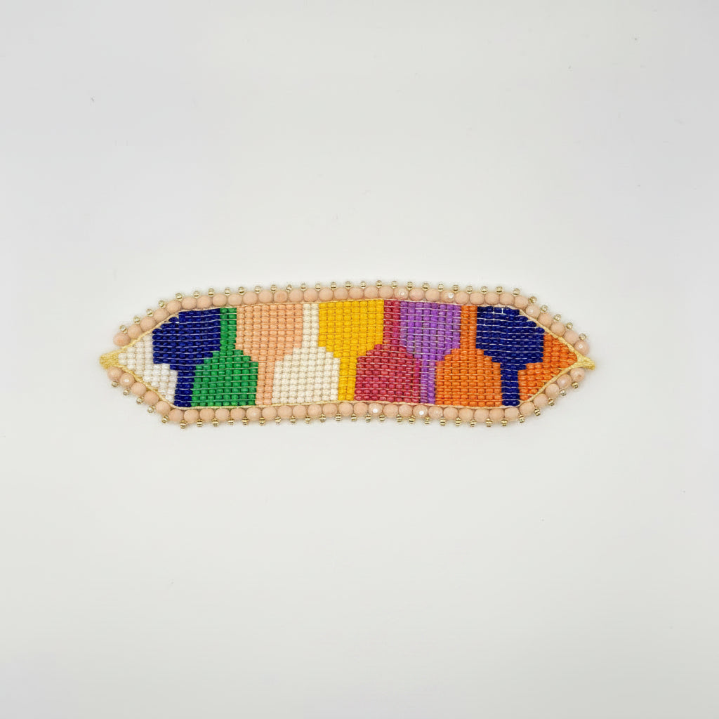 A handmade beaded hair claw clip by LOLA bloom, featuring a colorful geometric pattern of blue, green, orange, yellow, pink, and purple beads, attached to a brown cardboard display card with a pink circular logo.