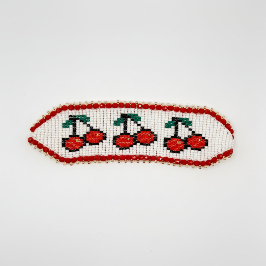 A handmade beaded hair clip featuring a white base decorated with three red cherries with green stems, bordered by red beads, with a brown cardboard backing and a pink paper insert.