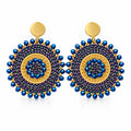 A pair of handmade circular beaded earrings with blue, purple, and gold concentric patterns, displayed on a white background.
