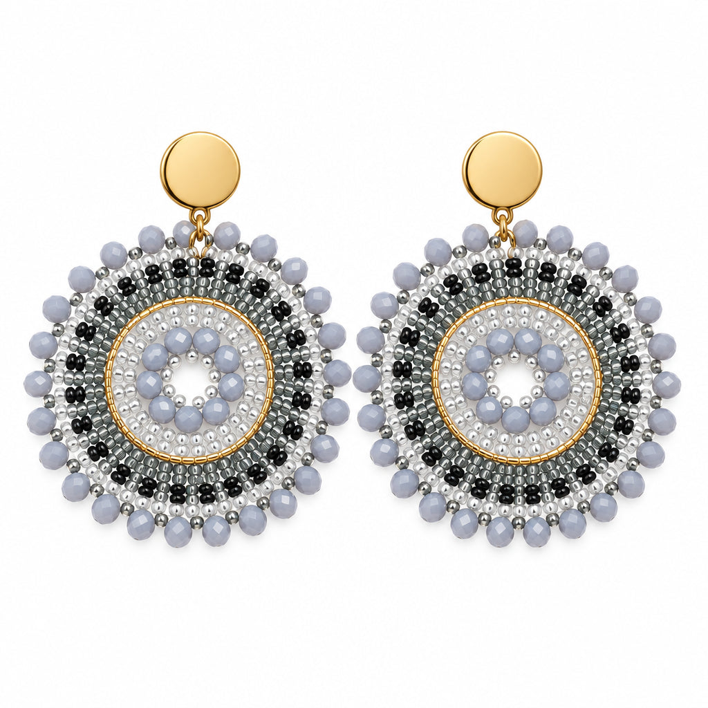 A pair of bohemian-style dangle earrings with circular silver and black beaded patterns, gold-toned posts, and white crystal centers, displayed on a white jewelry stand.
