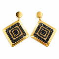 A pair of gold-toned geometric drop earrings with black beaded concentric square patterns, displayed on a clear card against a white bust form.