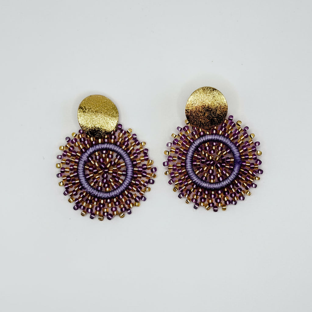 A pair of handmade beaded earrings featuring purple and gold seed beads in a circular sunburst pattern with a textured gold disc at the top, displayed on a clean white background.