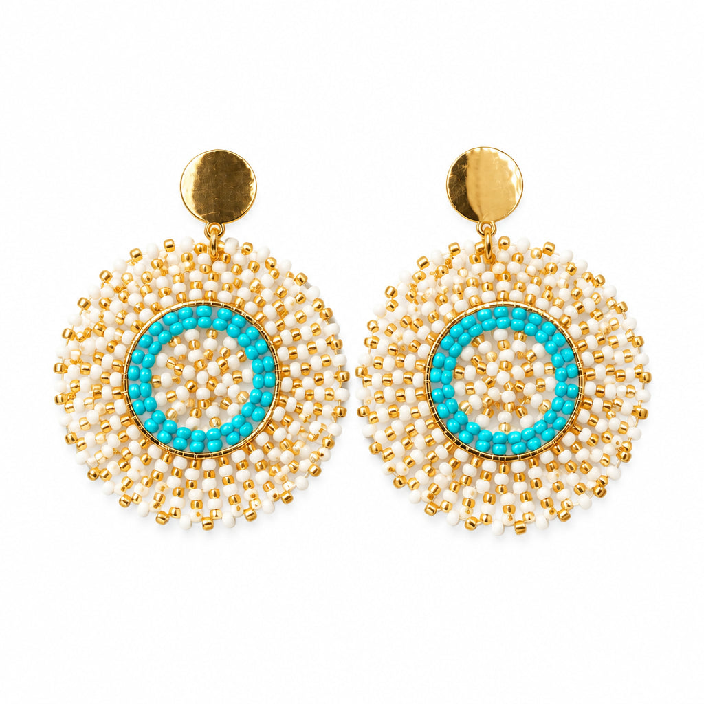 A pair of gold-toned statement earrings with a turquoise blue circular center and white bead fringe, displayed on a clear plastic card against a white background.