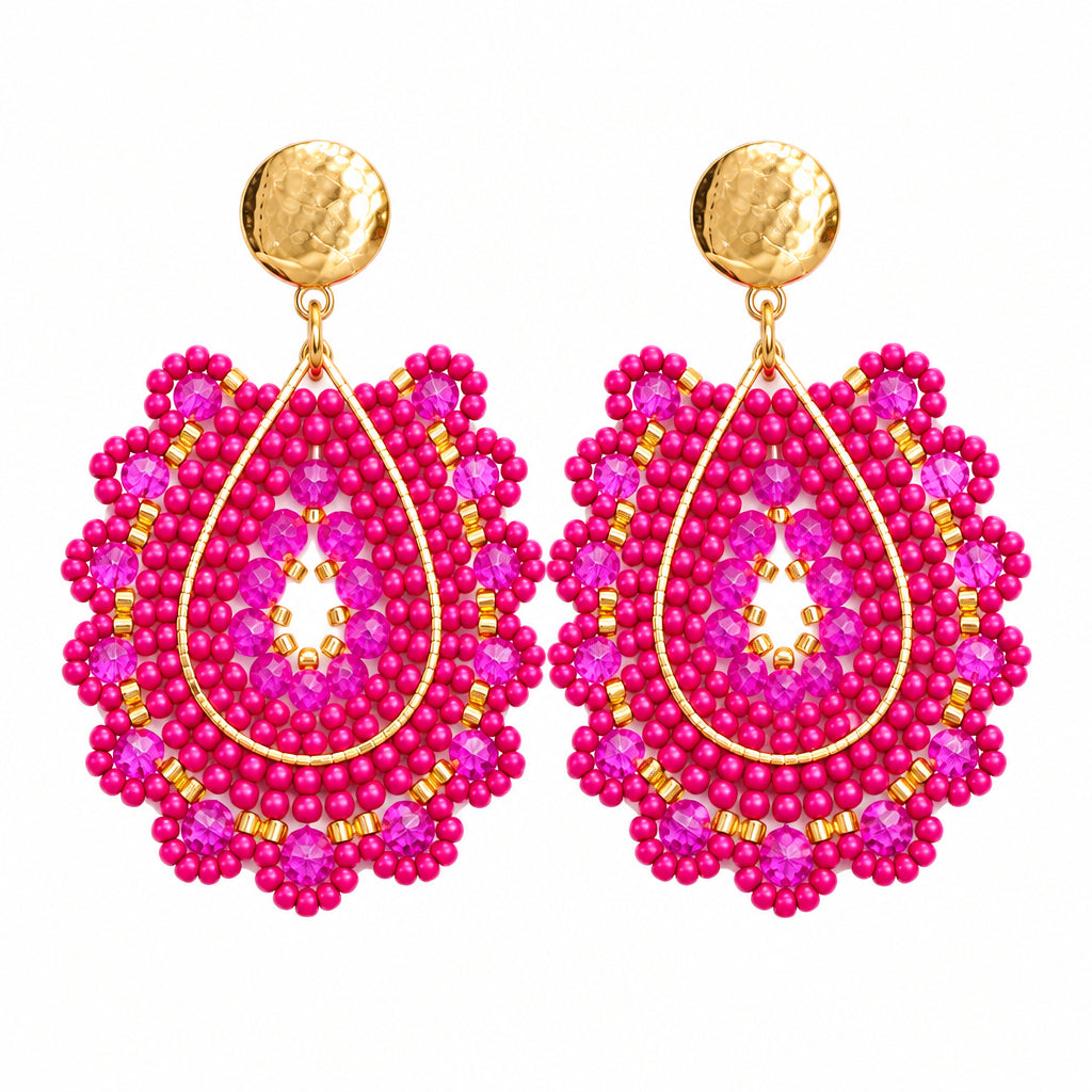 A pair of statement dangle earrings featuring intricate beaded teardrop shapes in vibrant magenta pink and gold, with hammered gold studs at the top, set against a solid light gray background.
