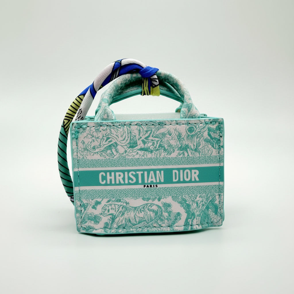 A Christian Dior handbag in a turquoise and white toile pattern, featuring a matching patterned scarf wrapped around its handles, set against a plain light gray background.