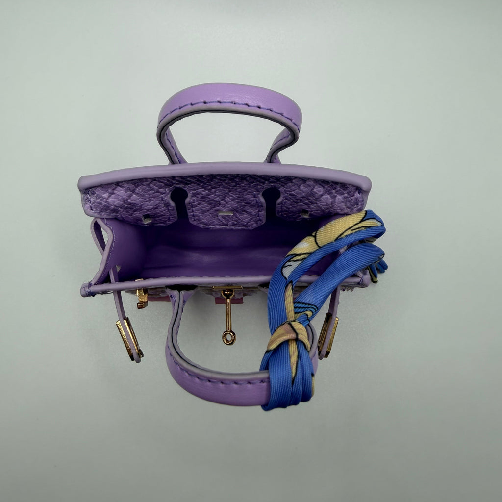 A small purple leather handbag shown open from above, with a purple snakeskin-patterned interior lining, gold-tone hardware, and a blue and beige patterned ribbon tied around the handle, set against a plain light gray background.