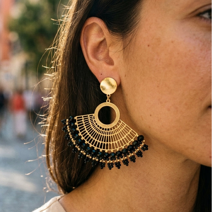 A close-up lifestyle shot of a woman wearing a pair of large gold and black fan-shaped dangle earrings, set against a blurred cobblestone street background.