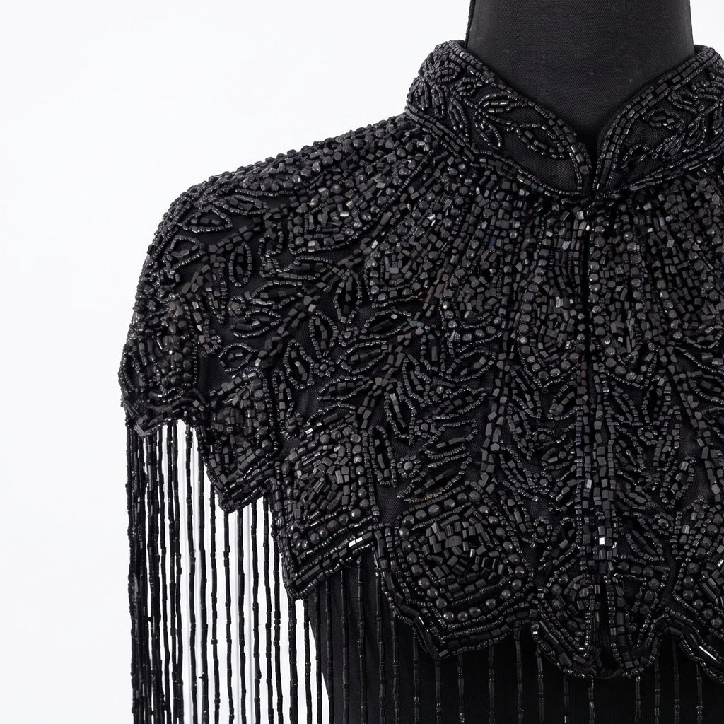 A black beaded high-neck shrug or cape with intricate beadwork and long hanging fringe, displayed on a black mannequin torso against a white background.