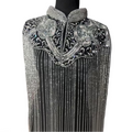 A close-up of a black embellished garment displayed on a mannequin, featuring heavy silver beadwork, sequins, floral appliqué, and long vertical fringe on the front.