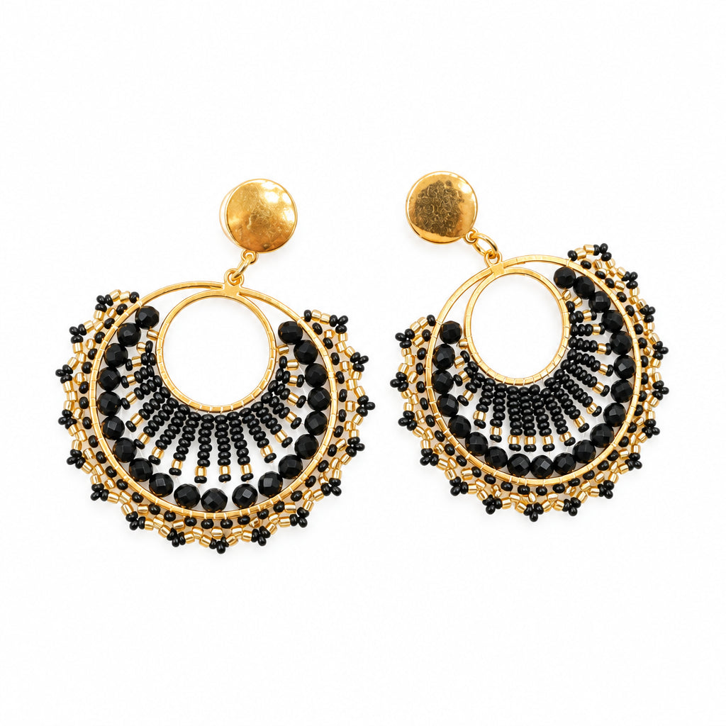 A pair of gold-toned statement earrings with black beaded fringe and white circular inlays, displayed on a clear card against a white background.