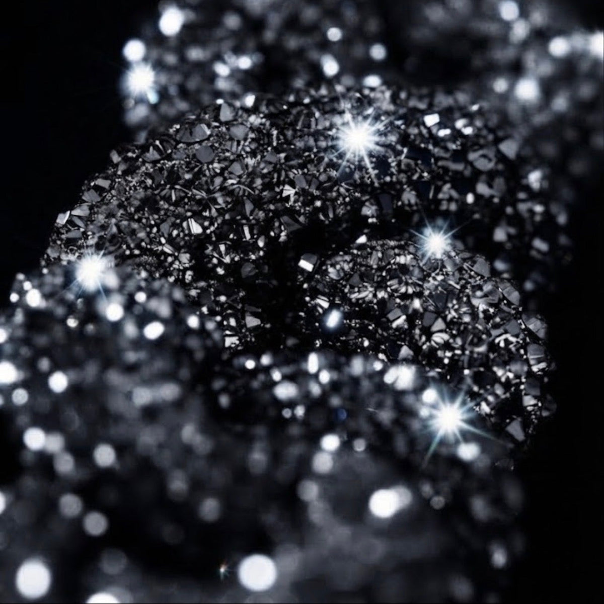 A close-up of sparkling black and silver glitter or crushed crystal texture against a black background, with bright reflective highlights.